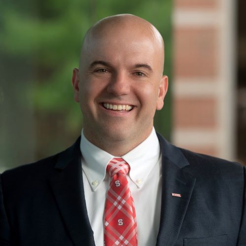 bald man wearing a black suit and an NC State red tie