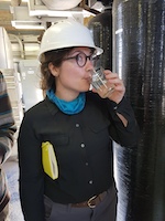Woman in hard hat drinking out of a beaker inside a lab.