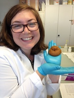 Woman in lab coat and glasses smiling while holding synthetic poop.