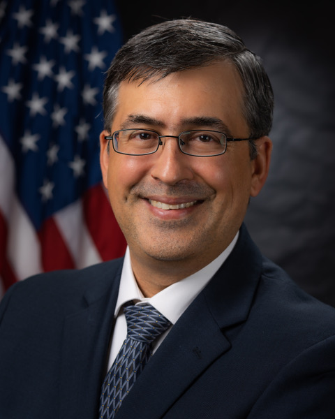 man with dark hair and glasses smiling in front of an American flag while wearing a suit and tie