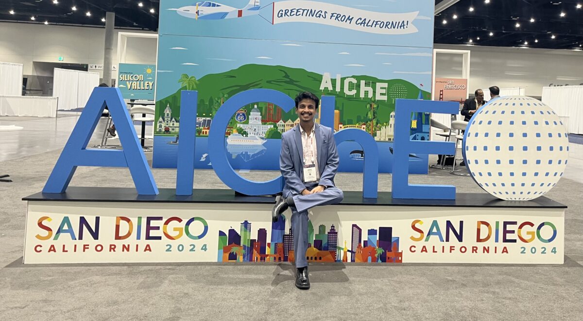 man in a suit sitting in front of the AIChE logo