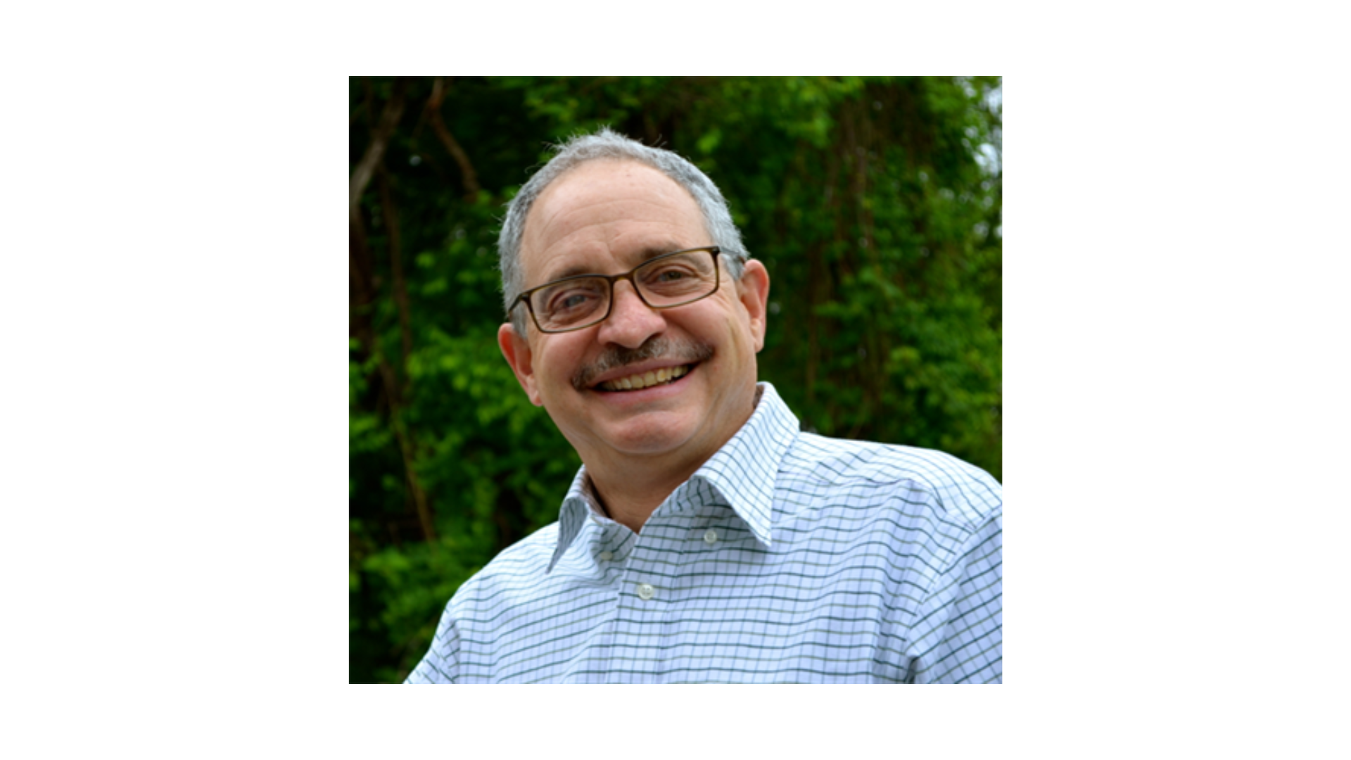 man with gray hair wearing a blue collared shirt and glasses smiling outside