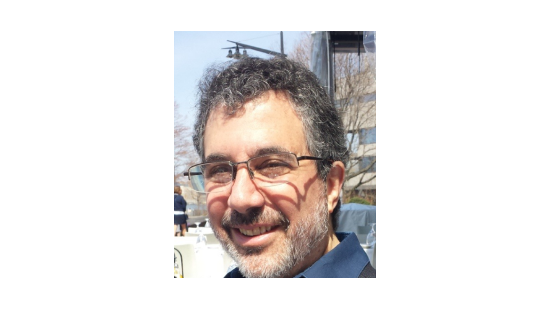 man with gray hair and facial hair wearing glasses and smiling over his left shoulder