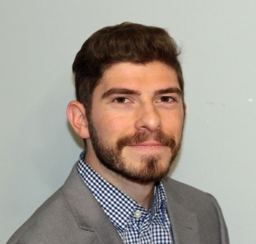 man with dark hair and facial hair wearing a gray blazer and smiling