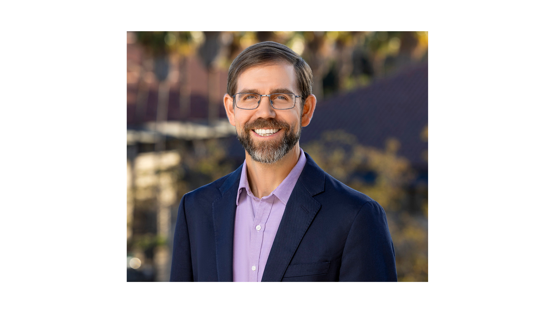 man with dark hair and a beard wearing glasses and a suit smiling outdoors