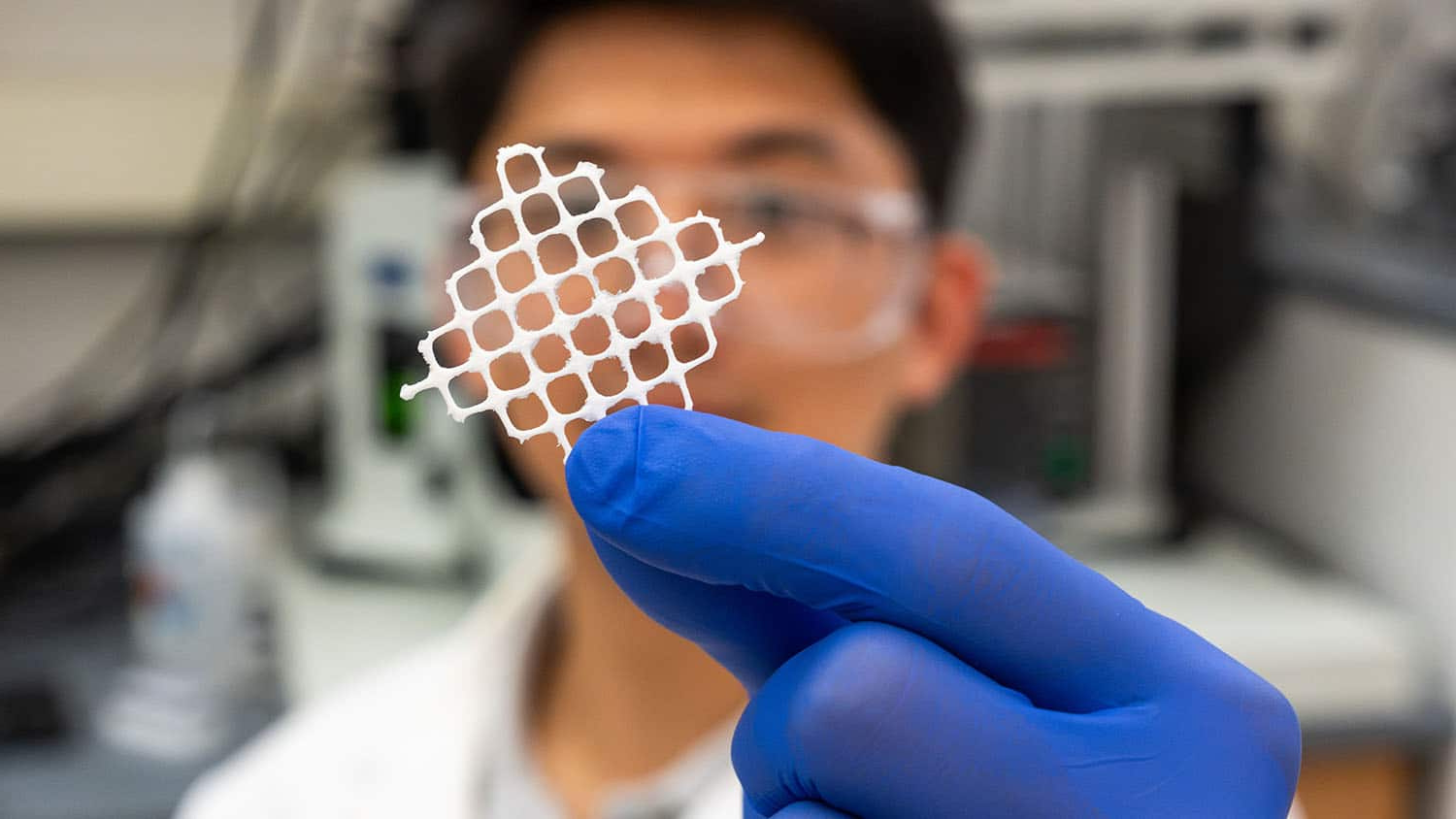 a researcher in a white lab coat and purple gloves holds a piece of white mesh up in front of the camera