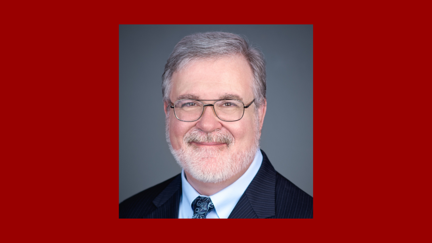 headshot of a man with short gray hair and close-shaven beard wearing a suit and glasses against a red background
